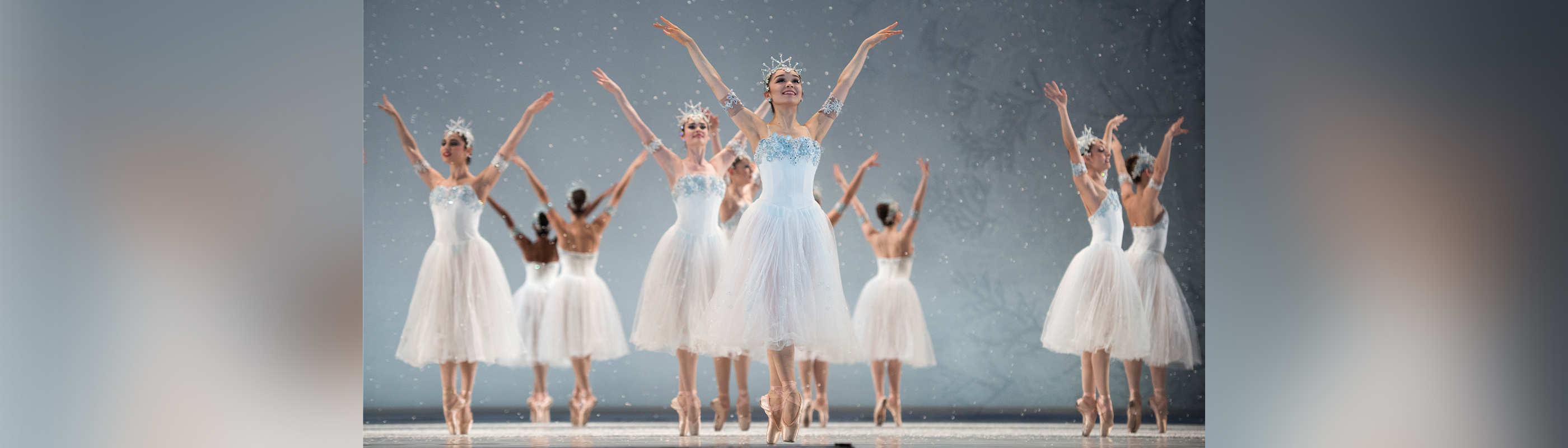 Snow falls as five pairs of Snowflakes stand on pointe with ankles crossed, facing away from each other, arms raised, palms out