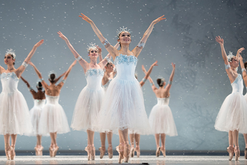 Snow falls as five pairs of Snowflakes stand on pointe with ankles crossed, facing away from each other, arms raised, palms out