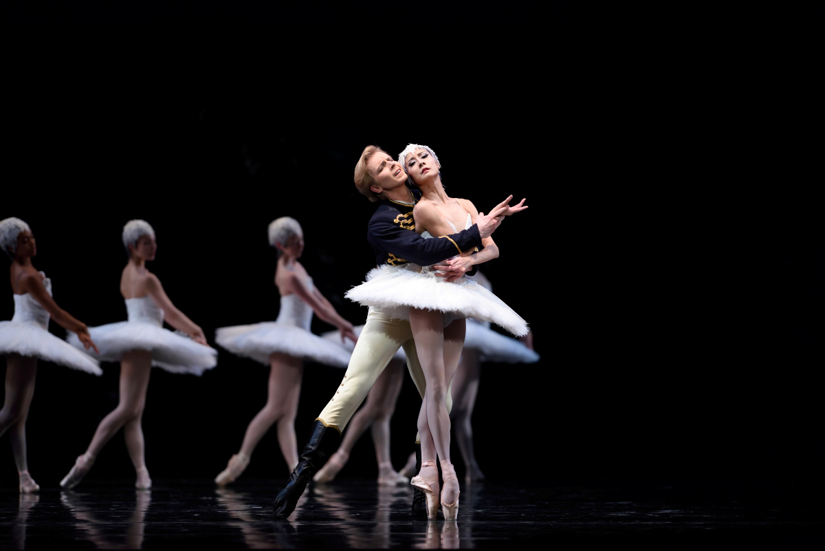 Before a line of swans facing left, right feet pointed behind, Siegfried caresses Odette as she balances on pointe, feet crossed