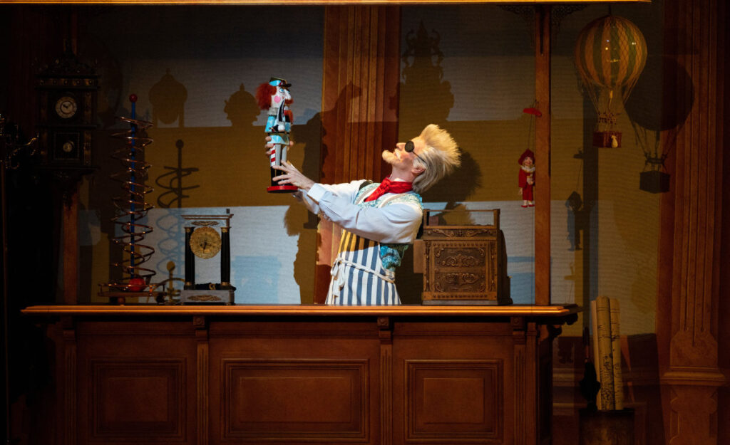 Drosselmeyer, standing at the counter of his shop, holding aloft a toy nutcracker.